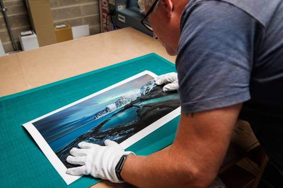 A person wearing white gloves and glasses examines a print of a wintry landscape with the Northern Lights in the sky above.