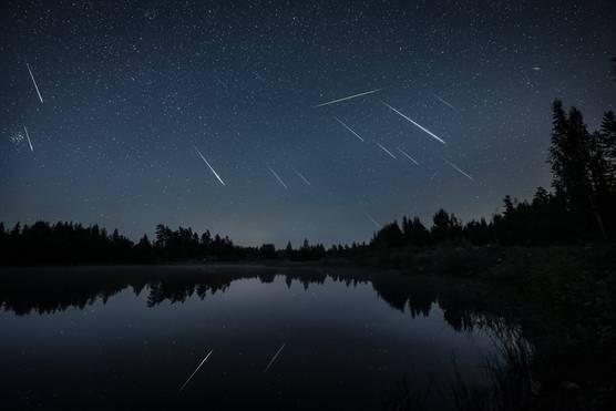 Meteors streak across a night sky, reflected in a still lake surrounded by silhouetted trees.