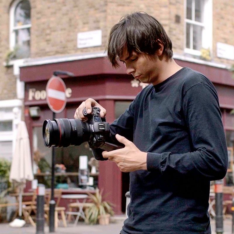 Молодой человек в черной одежде стоит на углу улицы, держа камеру Canon EOS R5 на уровне груди и глядя на ЖК-экран.
