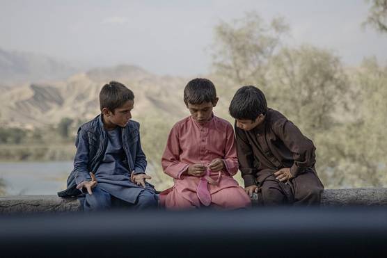 Three young Afghan boys deep in discussion sit on a wall in front of a large body of water. One of the boys is holding a bright pink sandal.
