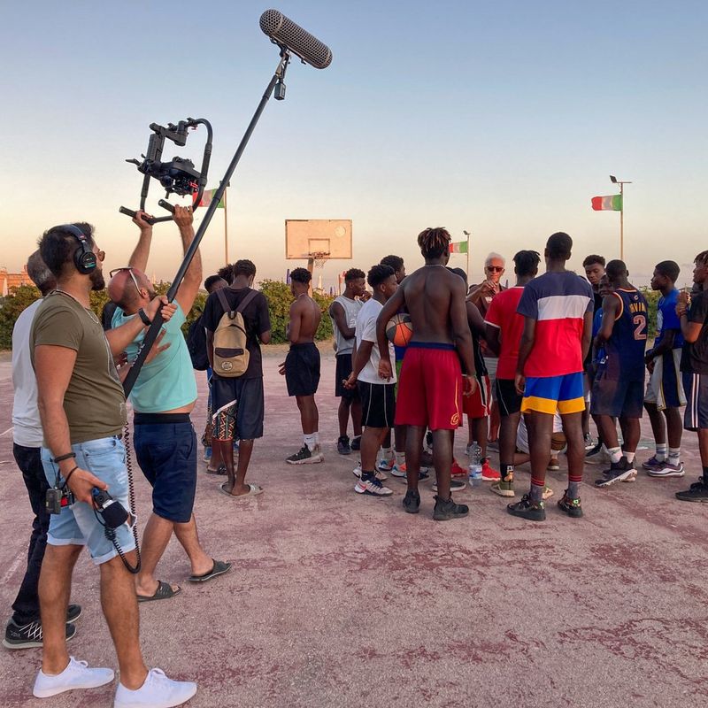 A group of basketball players gather together on a court, standing around their coach. To the side, three filmmakers stand with cameras and a microphone, filming them. 