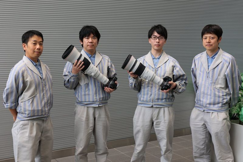 Four men dressed in the same grey and blue lab coats in front of a wall. Two of them are holding Canon super-telephoto lenses.