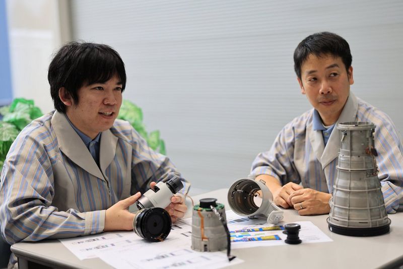 Two men dressed in striped lab coats sit at a desk. In front of them are parts of a Canon super-telephoto lens.