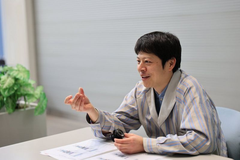 A man wearing a striped lab coat sits at a desk. He is gesticulating with one hand and holding part of a Canon lens in the other.
