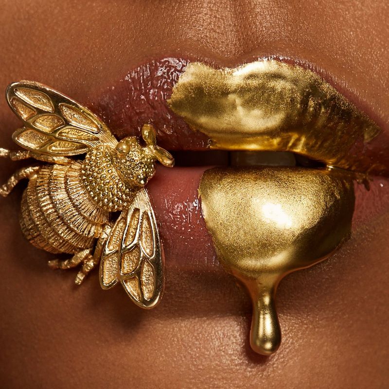 A gold metal bee sits on a female model's made-up lips that are dripping with gold paint.