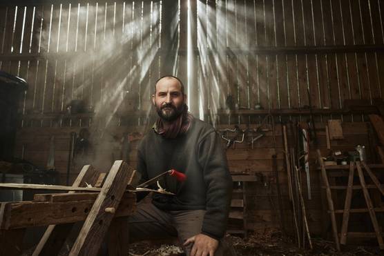 A bearded man sits in a barn surrounded by basketmaking tools, looking straight at the camera, in a portrait taken on a Canon EOS R5 C by Tom Barnes.