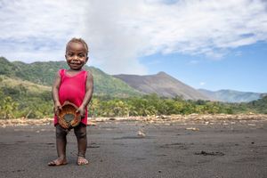 Фотография Уллы Лохманн, снятая на камеру Canon EOS R5, изображает ребенка в ярко-красном платье на фоне дымящегося вулкана
