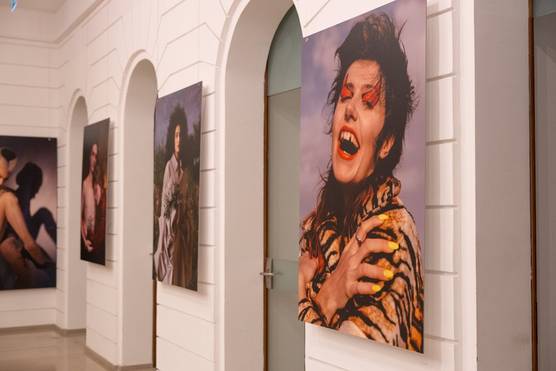 Four portraits on a gallery wall. The one closest to the camera shows an androgynous person wearing heavy make-up and laughing. 