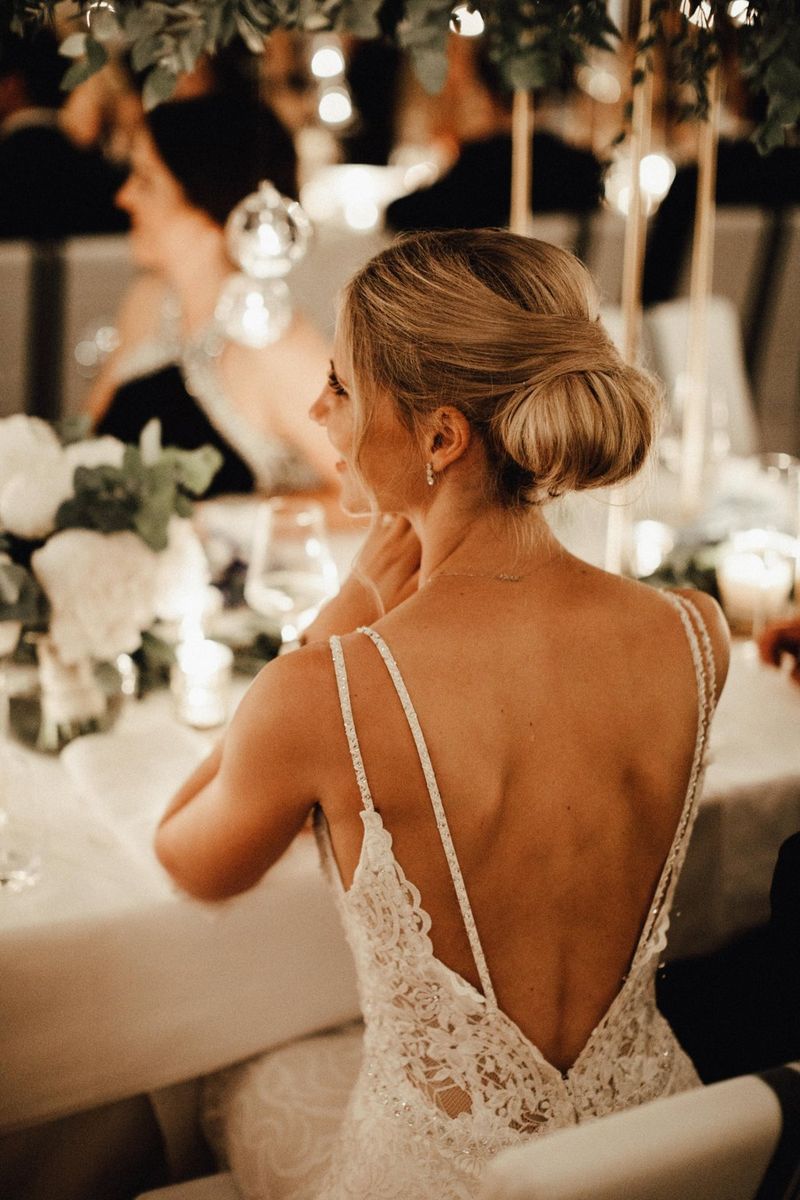 A portrait, shot from behind, of a bride sat at a decorated top table. Her wedding dress has a deep 'v' at the back. 