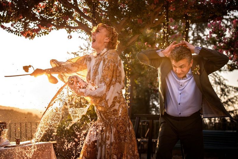 A bride opening a bottle of champagne with a sword in the late afternoon light. Her new husband stands by her side with his hands over his head.