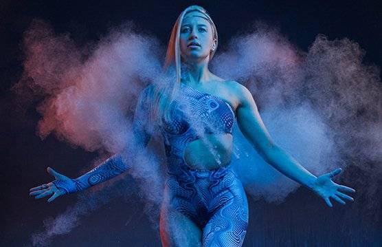 A predominantly blue-toned photo taken by James Musselwhite of a female model with arms stretched standing against a dark background as smoke engulfs her.