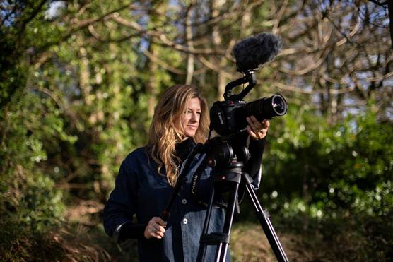 A woman with dark blonde hair films in a forest setting with a Canon camera on a tripod. 