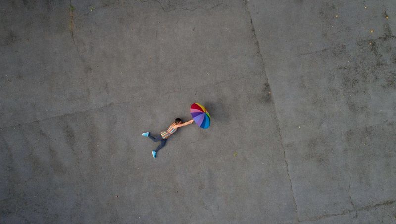 Shot from a drone high above, a woman lies on the floor holding a rainbow-striped umbrella.