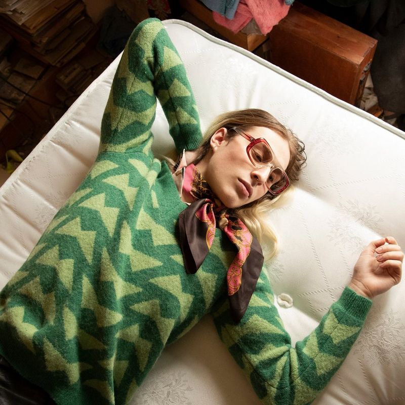 An androgynous long-haired model wearing a striped top and scarf lies on a bed.