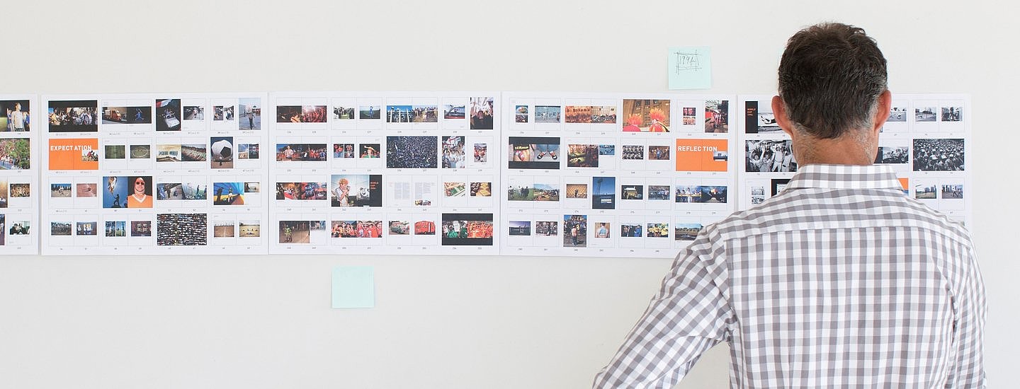 Man in a checked shirt with his back towards the camera stands with his hand on his hip as he stares at tiny images spread across a white wall.