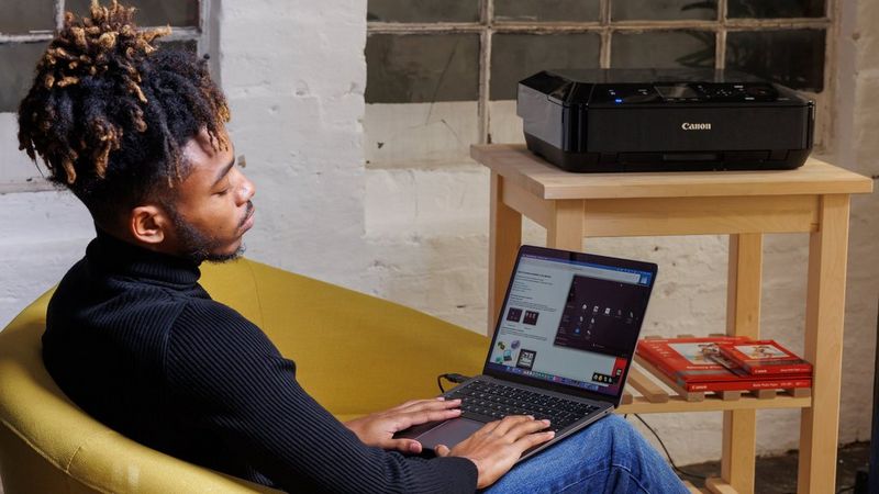 A black man sitting on a sofa with laptop on his lap with Quick Menu software showing on the screen. A black Canon printer and photo paper seen in the background on a table.