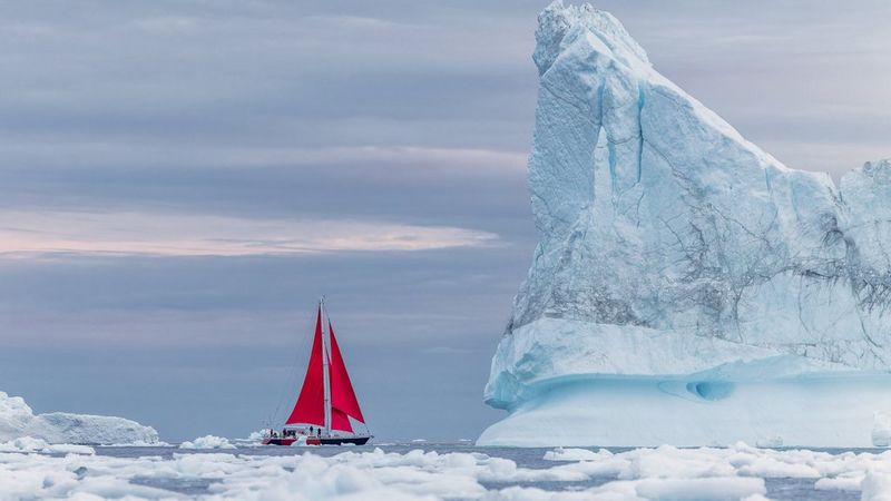 Um barco com velas vermelhas flutua nas águas geladas. À sua direita está um glaciar enorme e pontiagudo.