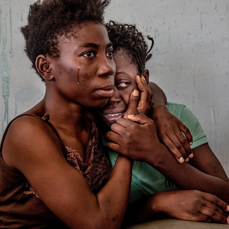 Two woman cling onto each other bravely. One has a tear rolling down her cheek, but looks ahead resolutely, while the other partially hides her face.