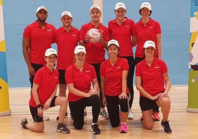 A netball team, posed for the camera with five people stood at the back and four on one knee at the front. They all wear red Canon branded polo shirts and white Canon branded baseball caps. The man stood at centre back holds a netball.