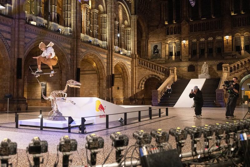 A female skateboarder jumps over a skeleton of a dinosaur from a ramp, in the Nature History Museum. Two male camera operators film the jump on Canon video cameras.