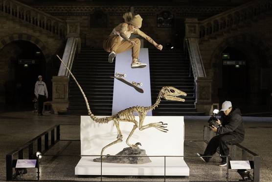 A female skateboarder jumps over the skeleton of a Velociraptor at the Natural History Museum in London, while being filmed by a person to the right.  
