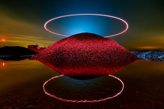 A mound of stone is illuminated by a halo of red light above it, which is also reflected in the water at its base.