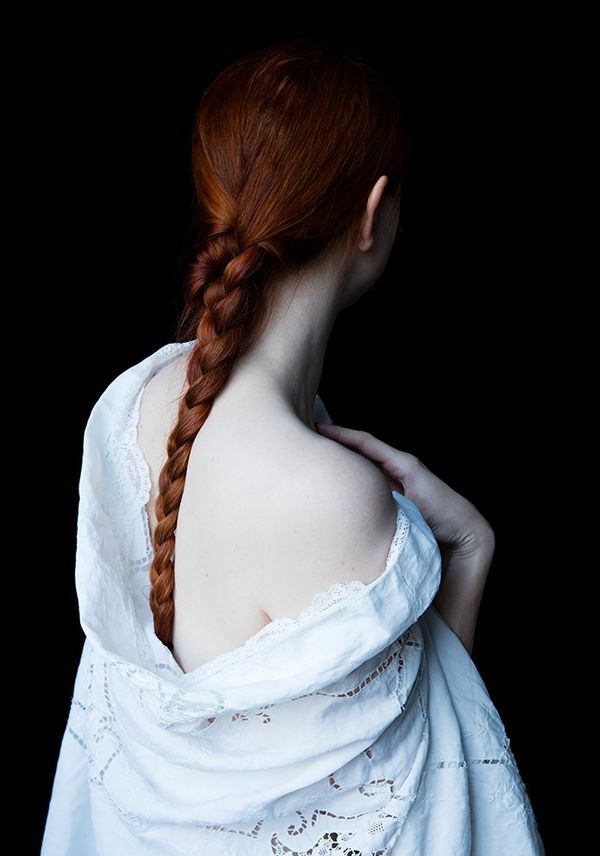 A woman with red hair tied in a plait faces away from the camera. She wears a low-backed dress that shows off her milky white skin.