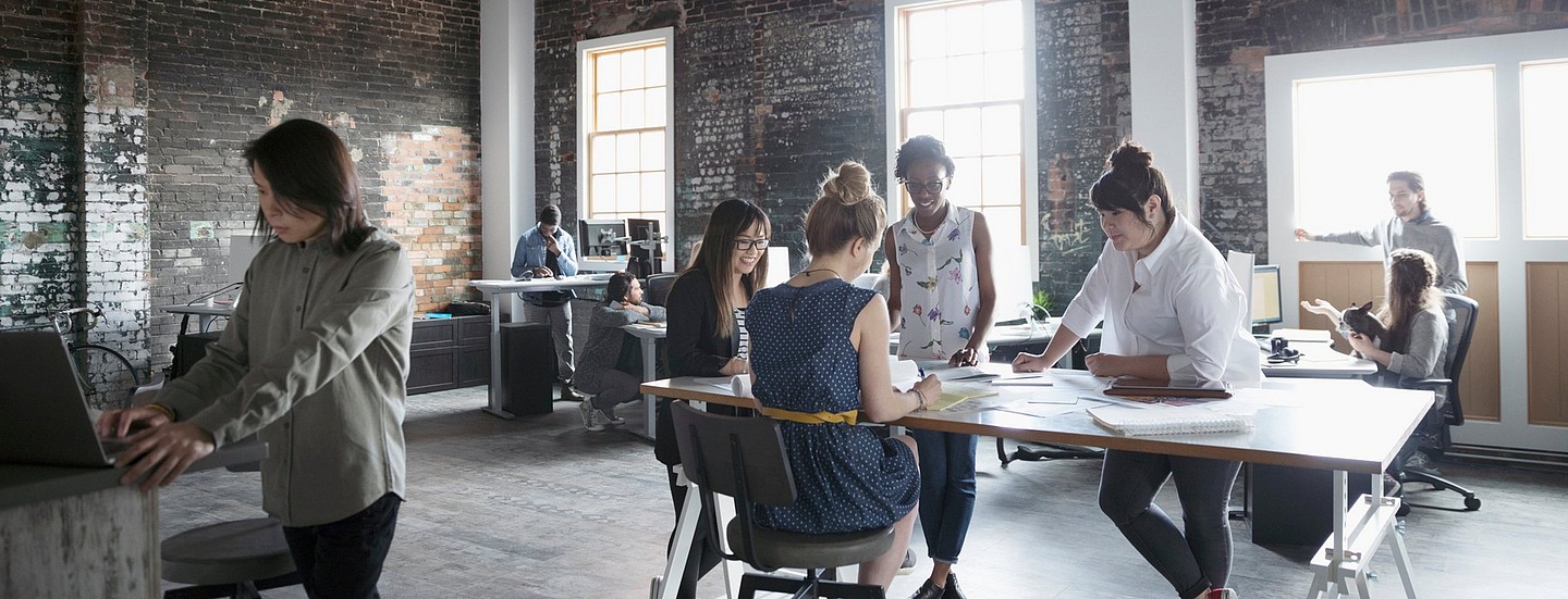 Casual exposed-brick office environment, with one group crowded round a table and others working at laptops and PCs.