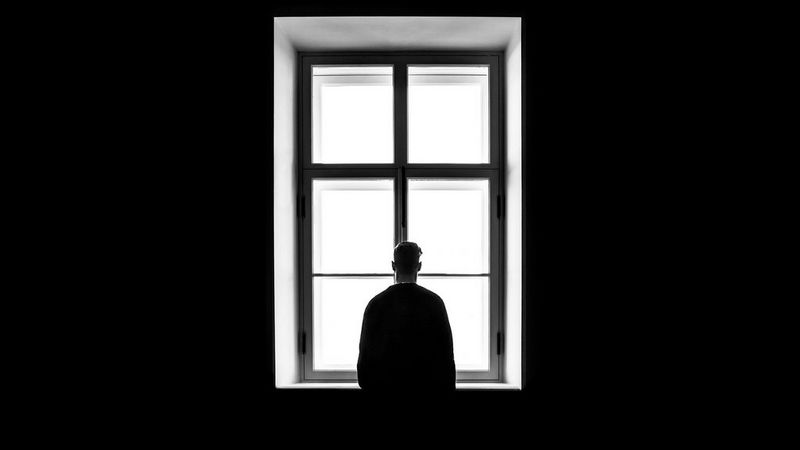 A black and white photograph of a back of a man, silhouetted, stood in front of at the bright white light of window. On either side, the walls are black.