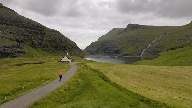 A person walks down a road towards a hut with a lake next to it as a waterfall flows down a hill in the Faroe Islands. Taken with a Canon RF 10-20mm F4L IS STM lens by Joel Santos.