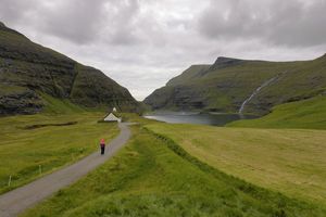 Eine Person läuft auf einer Straße in Richtung einer Hütte, neben der sich ein See befindet. Auf einem Hügel der Färöer fließt ein Wasserfall nach unten. Aufgenommen von Joel Santos mit einem Canon RF 10-20mm F4L IS STM Objektiv.