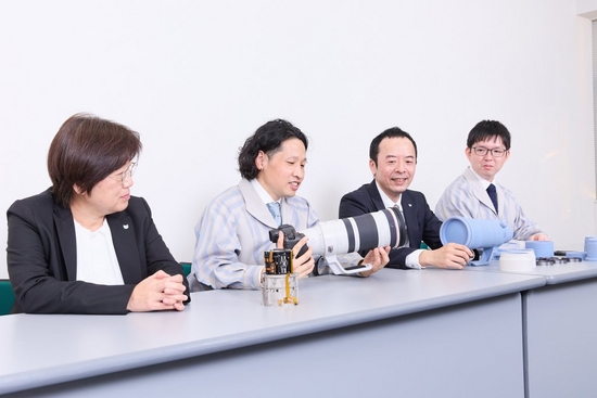 Four of the developers of the RF 100-300MM F2.8L IS USM sit at a table, one of them holding a camera with the lens attached.