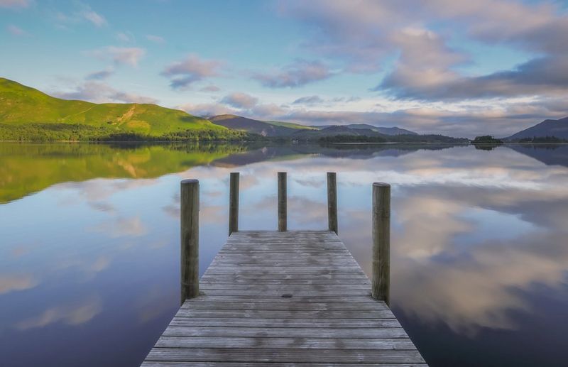 A wooden jetty over a lake surrounded by mountains. The clouds overhead are reflected on the lake's surface.