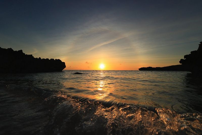 Waves break on the shoreline as the sun sets in the background. The light has created a halo effect around the sun, which sits between the silhouetted coastline.