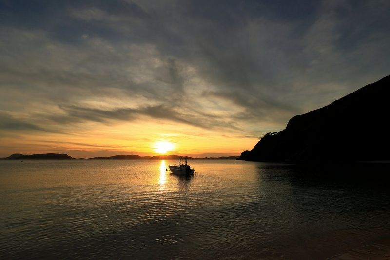 A small boat, lit by the glow of the setting sun in the background, on a calm body of water. The coastline in the foreground leads the viewer's eye around the frame as it extends into the distance.