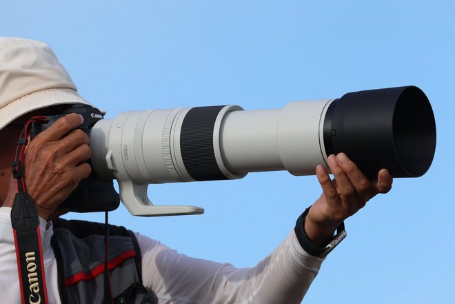 Un fotografo con cappello da sole tiene in mano una fotocamera Canon a cui è fissato un obiettivo Canon RF 200-800mm F6.3-9.0 IS USM con rivestimento bianco.