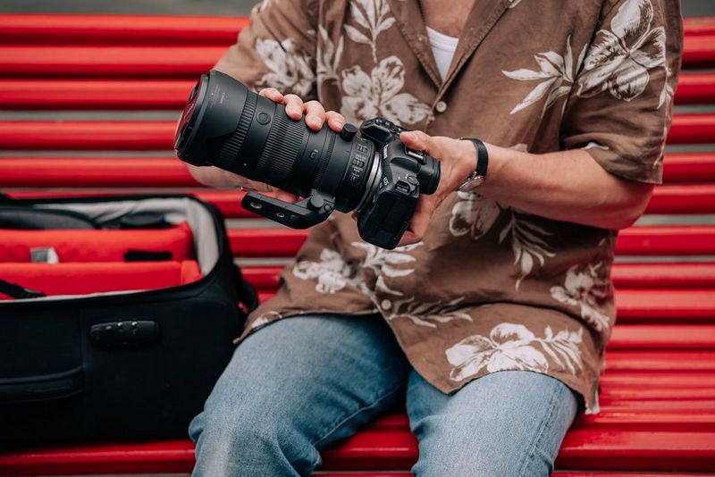 A user wearing a floral shirt and seated on a bright red wooden bench attaches a Canon RF 24-105mm F2.8L IS USM Z lens to a Canon camera.