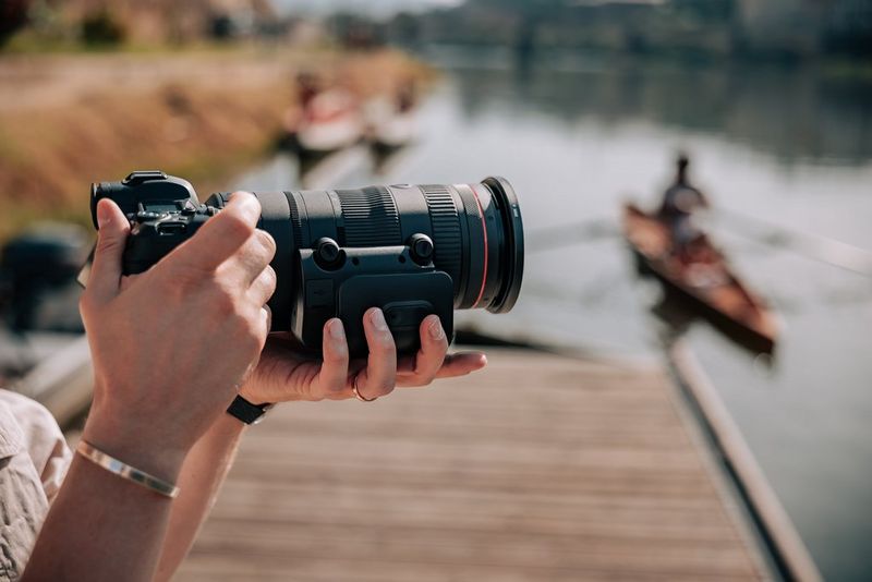 A user holds a Canon camera with a Canon RF 24-105mm F2.8L IS USM Z lens fitted with a Power Zoom Adapter. A rower on a river is visible in the background.