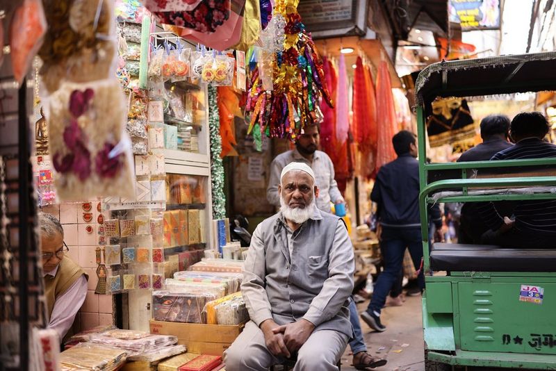 A wide-angle environmental portrait of a man with a white beard sitting by a roadside stall with colourful goods on display, taken with a Canon RF 35mm F1.4L VCM lens.