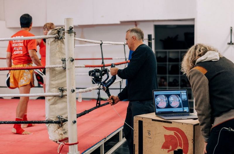 A man filming two martial arts practitioners in a ring with a Canon EOS R5 and a Canon RF 5.2mm F2.8L Dual Fisheye lens. A woman behind him is viewing the footage on a laptop.