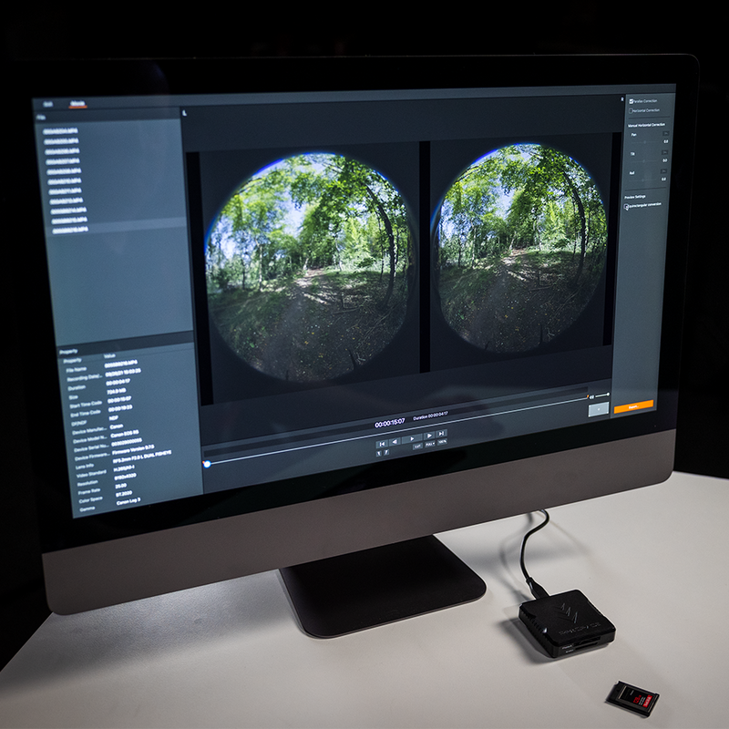 A computer monitor displaying two circular images of a forest path, captured with a VR lens. The monitor is placed on a white desk, and a memory card reader is connected to it.