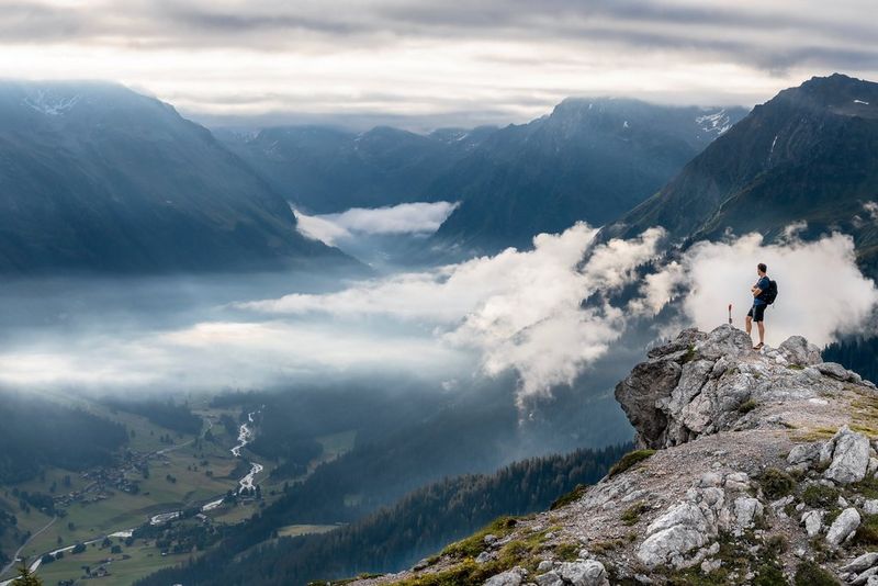 A lone walker stands on the edge of a rocky outcrop, framed against low clouds in a mountainous landscape with a steep-sided valley below.