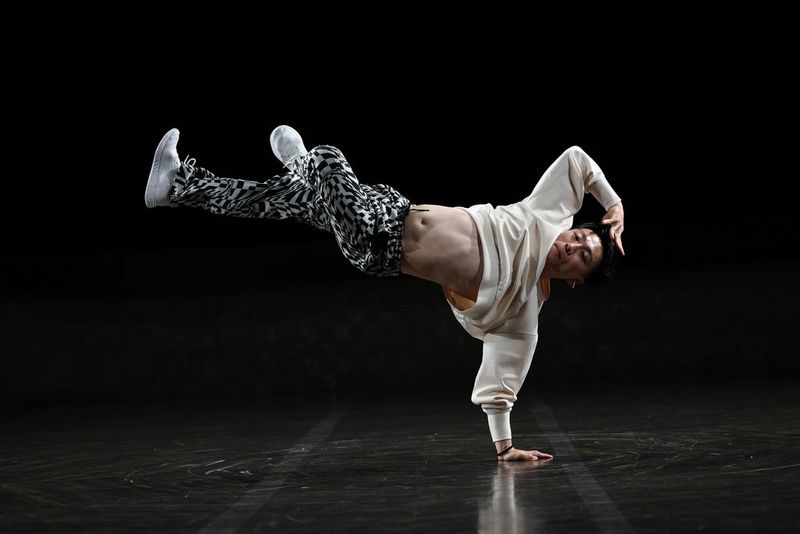 A portrait of an athletic dancer in a white sweatshirt and patterned black-and-white trousers, captured in mid-move, his body horizontal and supported on one hand, taken on a Canon RF 70-200mm F2.8 L IS USM Z lens.