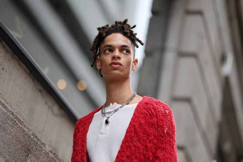 A low-angle portrait of a man in a red cardigan, standing in front of an out of focus concrete wall with bokeh lights.