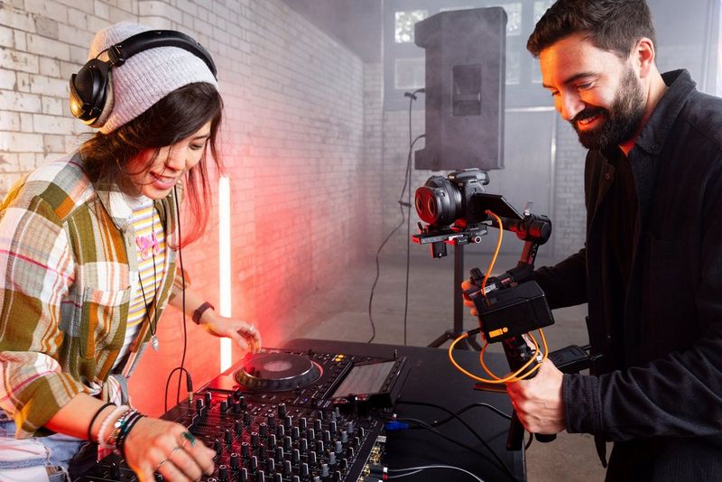  A woman wearing a beanie and plaid shirt is DJing with turntables and a mixer. Next to her, a photographer operates a camera with a Canon RF-S 3.9mm F3.5 STM DUAL FISHEYE lens attached, mounted on a gimbal.