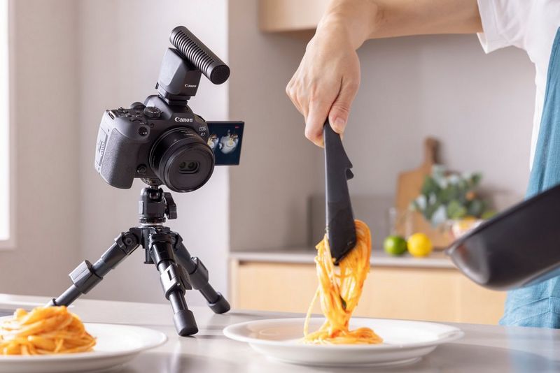 A Canon EOS R7 camera with a Canon RF-S 7.8MM F4 STM lens is mounted on a tripod, positioned on a white kitchen counter, aimed at a plate of spaghetti as someone uses tongs to serve it.