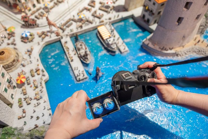  Two hands holding a Canon camera over a miniature harbour scene with blue water, boats and buildings. The camera’s screen displays circular stereoscopic  images created from the Canon RF-S 7.8MM F4 STM lens.