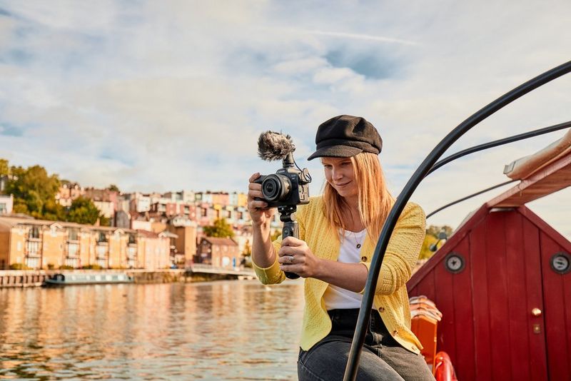 شابة تجلس على حافة قارب وهي تحمل كاميرا EOS R6 من Canon على مقبض حامل ثلاثي القوائم ويوجد في الخلفية نهر وصفّ من المنازل.