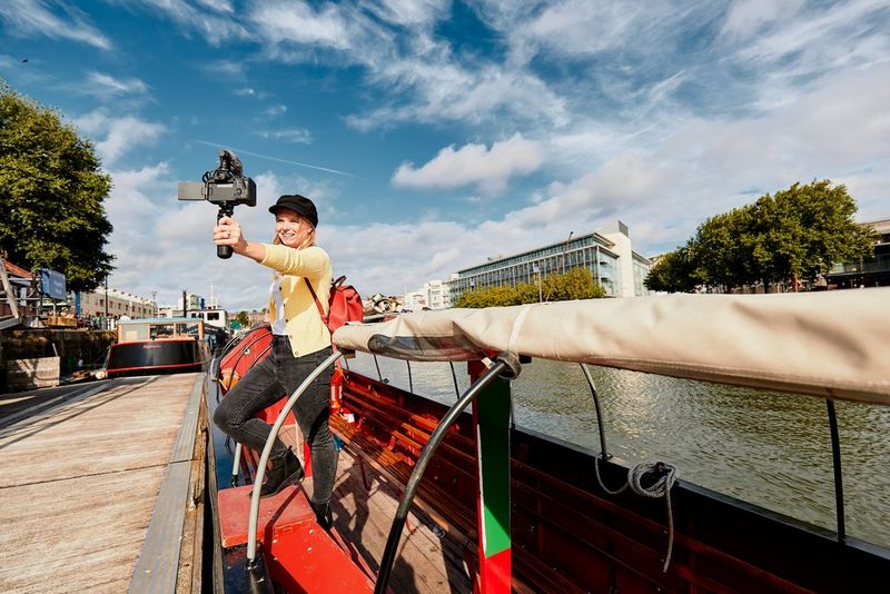 A young woman filming herself on a Canon EOS R6 with a Canon RF 16mm F2.8 STM lens stands on the edge of a boat moored next to a wooden jetty.