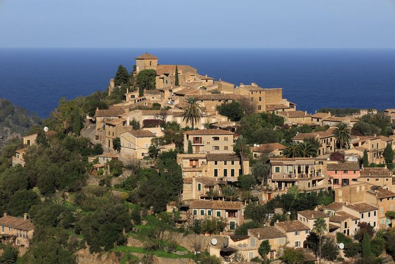 A landscape image of a small town built into the side of a hill. The sea and sky, both a vivid blue, can be seen in the background.